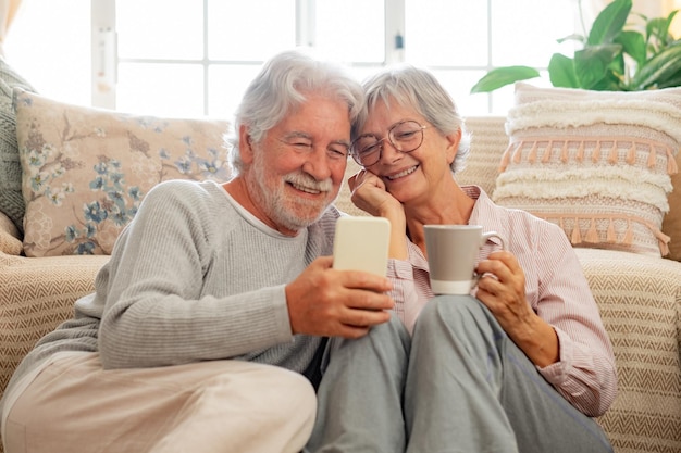 Liebevolles altes Senioren-Familienpaar, das zu Hause auf dem Boden sitzt und gemeinsam auf das Smartphone schaut