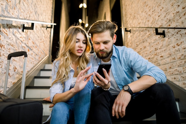 Liebevolles junges Paar in Freizeitkleidung mit Handy, auf der Treppe sitzend, im stilvollen Loft-Hotelflur