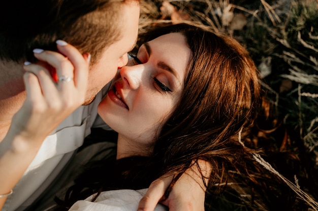 Liebevolles Paar, das im Sommer bei Sonnenuntergang auf dem Gras liegt und küsst. Brünettes Mädchen in einem leichten Kleid. Liebesgeschichte.