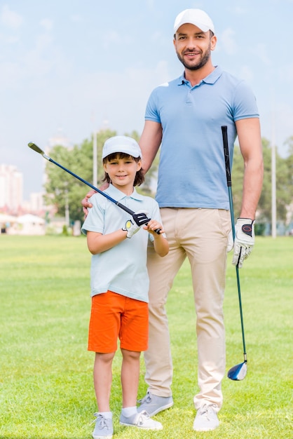 Lieblings-Familienspiel. Fröhlicher junger Mann, der seinen Sohn umarmt, während er auf dem Golfplatz steht