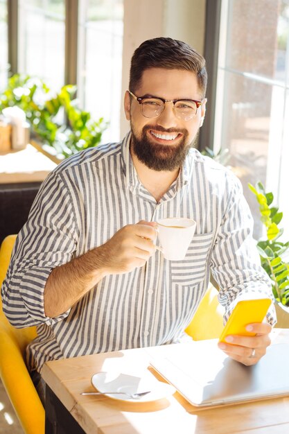 Lieblingsgetränk. Fröhlicher bärtiger Mann, der sein Smartphone hält, während er seinen Morgenkaffee im Café trinkt