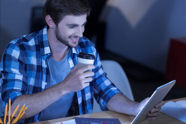 Lieblingsgetränk. Positiv freudig entzückter Mann, der Nachrichten liest und seinen Kaffee genießt, während er sich unterhält