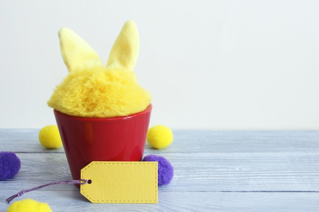 Liebres peludas amarillas del juguete en un pote de cerámica rojo en los tableros de madera grises, etiqueta de papel amarilla.