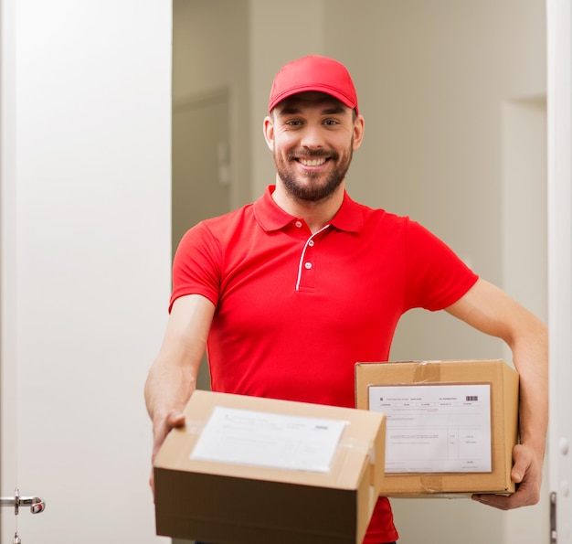 Liefer-, Post-, Personen- und Versandkonzept - glücklicher Mann in roter Uniform mit Paketkästen an offener Kundentür