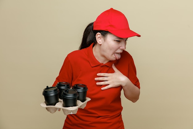 Foto lieferangestellte in roter mütze und leerer t-shirt-uniform mit kaffeetassen, die mit angewidertem gesichtsausdruck beiseite schauen und die zunge herausstrecken, die über braunem hintergrund steht