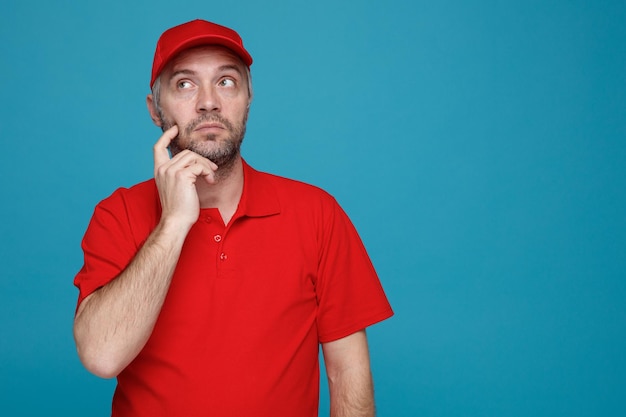 Lieferbote-Angestellter in der roten T-Shirt-Uniform der Kappe, die oben mit dem nachdenklichen Ausdruck denkt, der über blauem Hintergrund steht