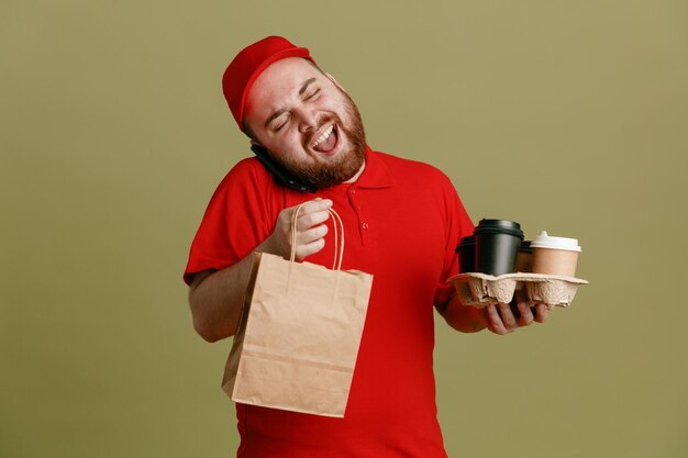 Lieferbote-Angestellter in der roten T-Shirt-Uniform, die Kaffeetassen und Papiertüten hält und am Handy spricht, glücklich und fröhlich lächelnd über grünem Hintergrund