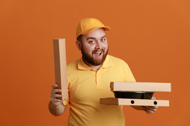 Lieferbote-Angestellter in gelber Kappen-T-Shirt-Uniform mit Lebensmittelbehältern und Pizzakartons, die glücklich und aufgeregt lächelnd in die Kamera schauen und breit über orangefarbenem Hintergrund stehen