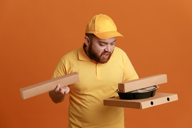 Lieferbote-Angestellter in gelber Kappen-T-Shirt-Uniform mit Lebensmittelbehältern und Pizzaschachtel, die verwirrt und unzufrieden über orangefarbenem Hintergrund stehen