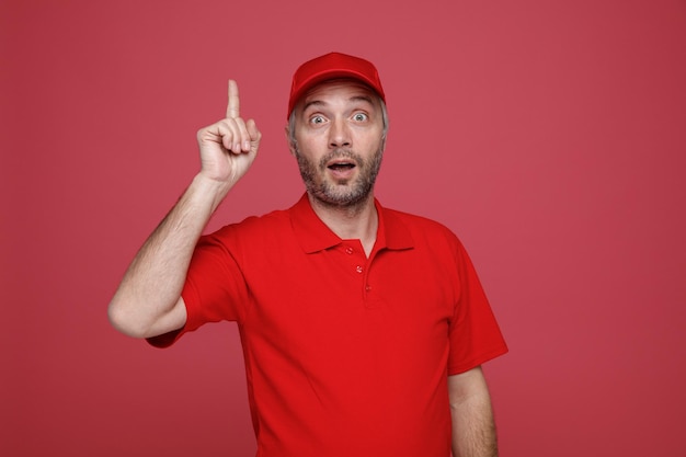 Lieferbote-Angestellter in roter Kappe, leerem T-Shirt, einheitlichem Blick in die Kamera, überrascht, Zeigefinger zu zeigen, der eine großartige Idee hat, über rotem Hintergrund zu stehen