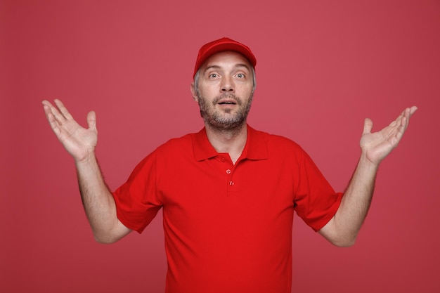 Lieferbote-Angestellter in roter Kappe, leerem T-Shirt, Uniform, Blick in die Kamera, verwirrt, die Arme hebend, mit Zweifeln, die über rotem Hintergrund stehen