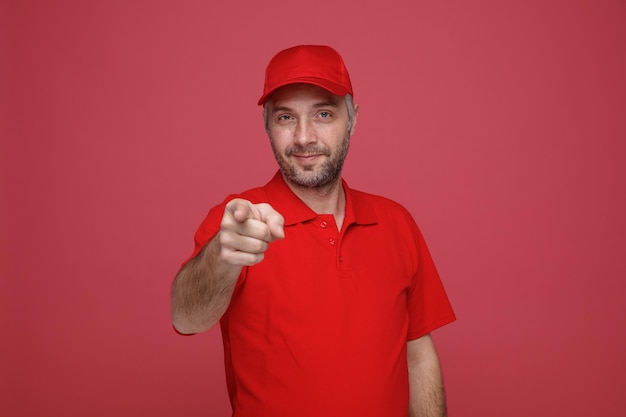 Lieferbote-Angestellter in roter Kappe, leeres T-Shirt, einheitliches Lächeln, freundlich, mit dem Zeigefinger auf die Kamera zeigend, die über rotem Hintergrund steht