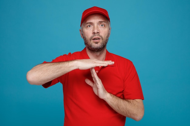 Lieferbote-Angestellter in roter Kappe, leeres T-Shirt, Uniform, der verwirrt in die Kamera schaut und eine Auszeit-Geste mit Händen macht, die über blauem Hintergrund stehen