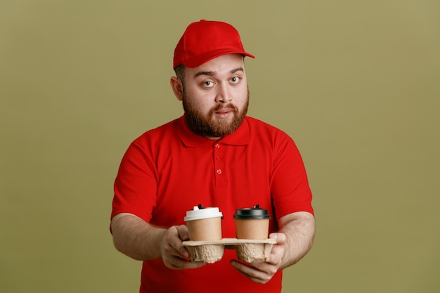 Lieferbote-Angestellter in roter Kappen-T-Shirt-Uniform mit Kaffeetassen, die mit selbstbewusstem Ausdruck auf grünem Hintergrund in die Kamera schauen