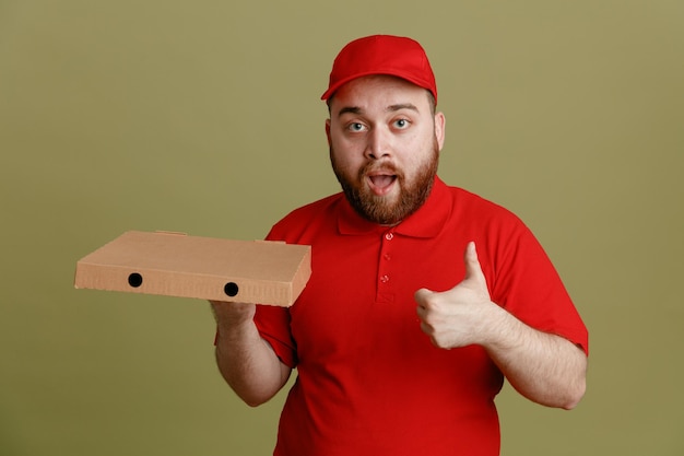 Lieferbote-Angestellter in roter Kappen-T-Shirt-Uniform mit Pizzakarton, der glücklich und positiv in die Kamera schaut und den Daumen nach oben zeigt, der über grünem Hintergrund steht