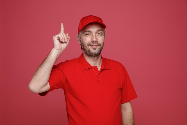 Lieferbote-Angestellter in roter T-Shirt-Uniform, die mit einem Lächeln auf dem Gesicht in die Kamera blickt und den Zeigefinger zeigt, der eine neue Idee hat, die über rotem Hintergrund steht