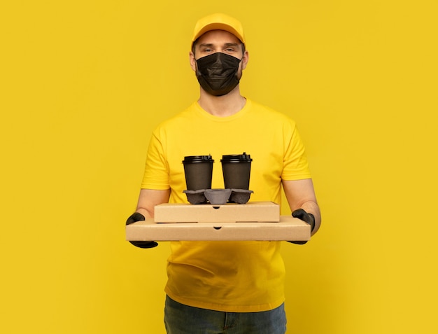 Lieferbote in gelber Mütze, T-Shirt Uniform, Maske, Handschuhe isoliert. Guy hält eine Tasse Kaffee zum Mitnehmen