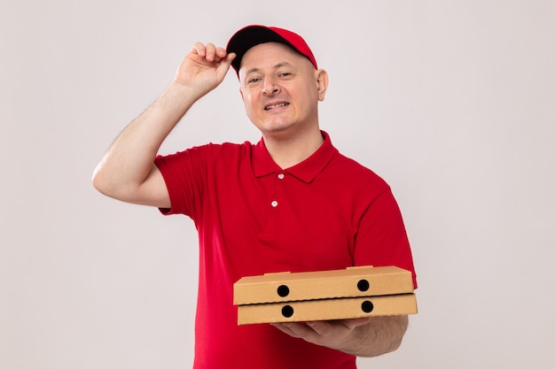 Lieferbote in roter Uniform und Mütze mit Pizzakartons, die glücklich und positiv lächelnd in die Kamera schaut und zuversichtlich auf weißem Hintergrund steht