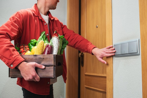 Lieferbote in Schutzmaske mit Papiertüte mit Essen im Eingang. Der Kurier steht vor der Tür und klingelt beim Kunden. Er hat eine Kiste mit frischem Gemüse und Obst mitgebracht