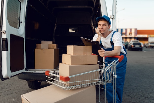 Lieferbote in Uniform hält Wagen mit Kisten am Auto mit Paketen, Lieferservice. Mann, der an den Papppaketen im Fahrzeug, im männlichen Liefer-, Kurier- oder Versandjob steht