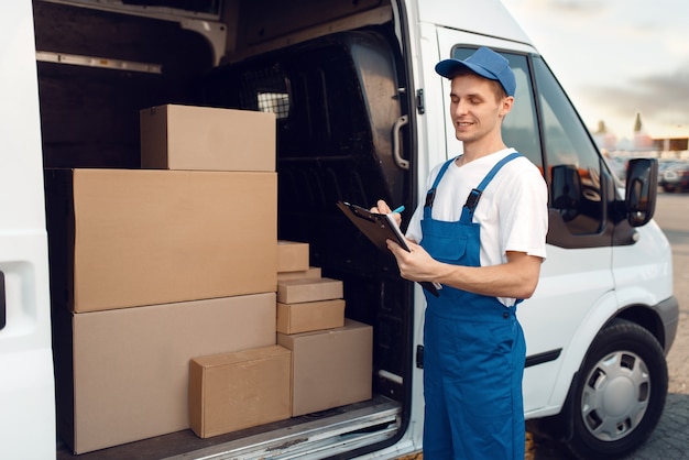 Foto lieferbote in uniform, kartons im auto, lieferservice. mann, der an den papppaketen im fahrzeug, im männlichen liefer-, kurier- oder versandjob steht