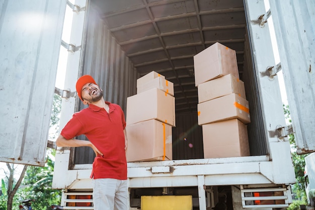 Lieferbote mit Rückenschmerzen beim Arbeiten mit Kartons hinter Container-LKW als Hintergrund