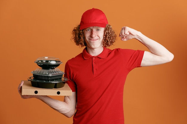 Lieferbote-Mitarbeiter in roter Kappe, leerem T-Shirt, Uniform, die Pizzakarton-Lebensmittelbehälter hält, die geballte Faust heben, glücklich und selbstbewusst lächelnd über orangefarbenem Hintergrund stehen