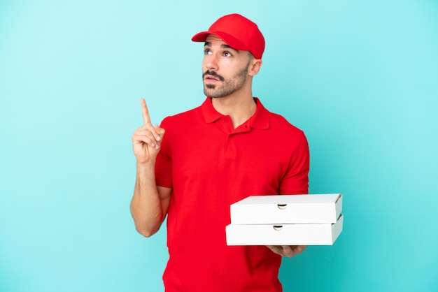 Lieferung kaukasischer mann, der pizzaschachteln isoliert auf blauem hintergrund aufhebt und eine idee denkt, die mit dem finger nach oben zeigt