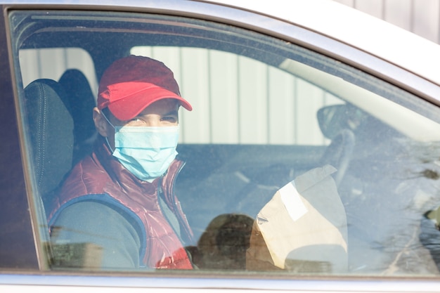 Lieferung mit dem Auto. Mann in Schutzmaske und medizinischen Handschuhen, der eine Papierschachtel hält. Lieferservice unter Quarantäne, Krankheitsausbruch, Coronavirus Covid-19-Pandemiebedingungen.