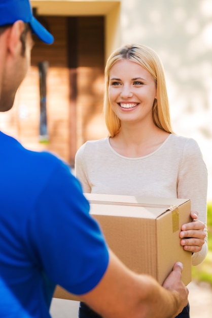 Lieferung von Hand zu Hand. Schöne junge Frau, die lächelt, während junger Lieferbote ihr einen Pappkarton gibt giving