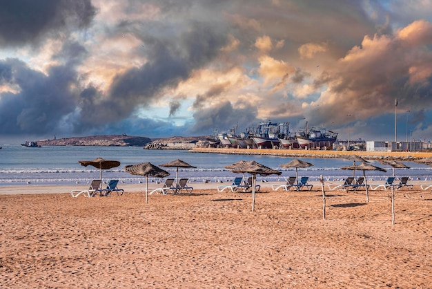 Liegestühle mit Sonnenschirmen in Reihe am Sandstrand an sonnigen Tagen gegen bewölktem Himmel, festgemachte Schiffe im Hintergrund