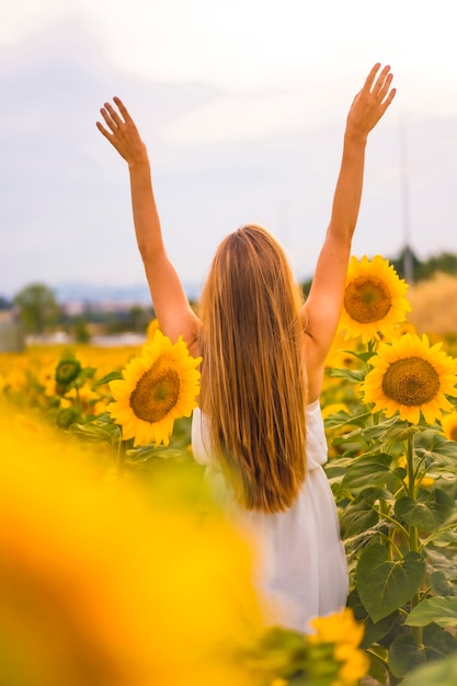 Lifestyle, una bella rubia caucásica en un campo de girasoles con un vestido blanco, con los brazos en alto