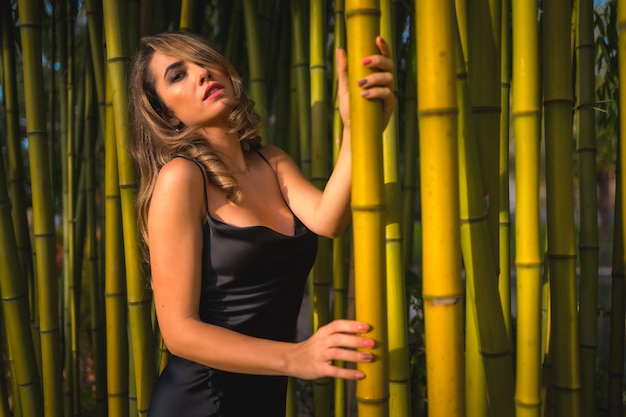 Lifestyle-blondes kaukasisches Mädchen des Lebensstils in einem glamourösen schwarzen Kleid im Park in der Natur thront auf einem schönen Bambusmodell, das nach links schaut