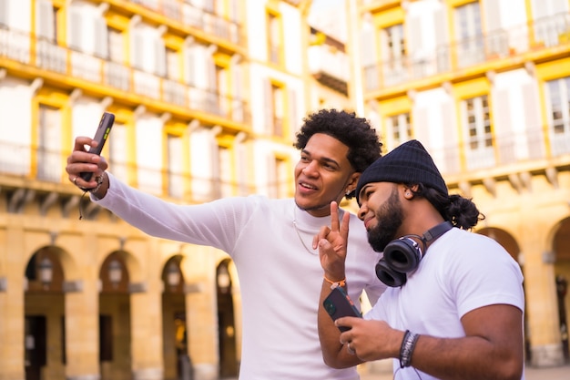 Lifestyle, dos amigos latinos negros tomándose una bonita selfie en la calle. Niño con pelo afro y niño con sombrero y auriculares