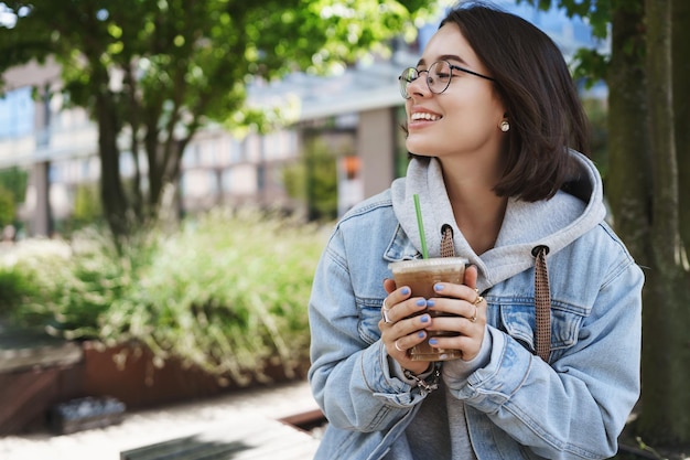 Lifestyle-Frauen- und Emotionskonzept Outdoor-Porträt eines glücklichen hübschen Mädchens mit Brille, das im Stadtpark steht und fröhlich lächelnd wegschaut und den Frühling beim Kaffeetrinken aus dem Lieblingscafé genießt