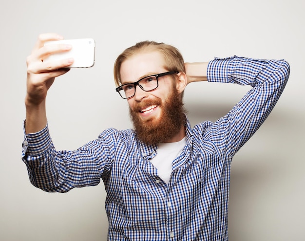 Lifestyle-Konzept Ein junger Mann mit Bart im Hemd, der ein Handy hält und ein Foto von sich macht, während er vor grauem Hintergrund steht