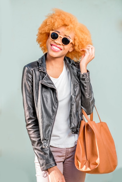 Lifestyle-Porträt einer afrikanischen Frau in Lederjacke und Sonnenbrille, die mit Tasche auf dem grünen Wandhintergrund steht