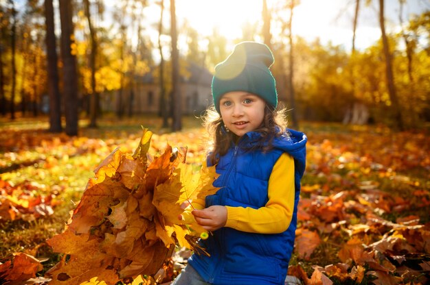 Lifestyle-Porträt. Schönes Kind, entzückendes Baby in leuchtend bunten warmen Kleidern, das im goldenen Herbstpark mit gesammeltem Strauß trocken gefallener Ahornblätter bei Sonnenuntergang mit fallenden Sonnenstrahlen sitzt