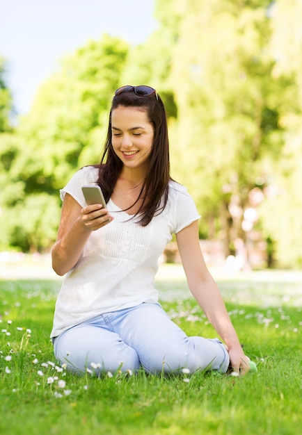 Lifestyle, Sommer, Urlaub, Technologie und People-Konzept - lächelndes junges Mädchen mit Smartphone, das auf Gras im Park sitzt