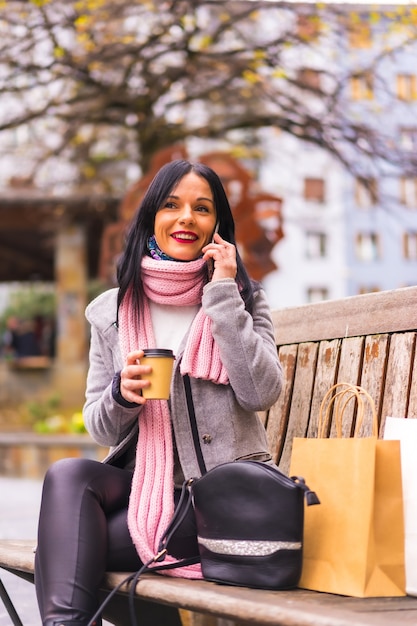 Lifestyle, uma menina morena caucasiana fazendo compras na cidade com sacos de papel e um café para viagem, sorrindo sentado no banco