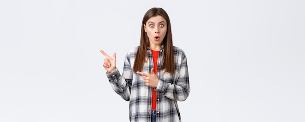 Lifestyle verschiedene emotionen freizeitaktivitäten konzept beeindrucktes weibliches kundenmädchen in lässig kariertem hemd sagt wow, spricht über nachrichten und zeigt mit dem finger auf weißen hintergrund