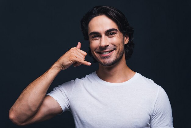 Liga para mim! Homem jovem e bonito sorrindo, olhando para a câmera e gesticulando para o celular perto do rosto em pé