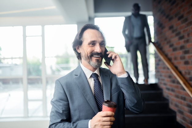 Foto ligando para um colega. homem grisalho segurando um café para viagem sorrindo enquanto liga para um colega