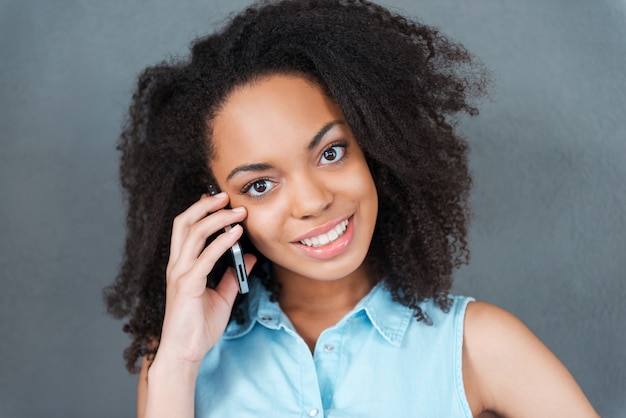 Ligando para você. Retrato de uma mulher jovem e atraente africana falando ao telefone e sorrindo em pé contra um fundo cinza