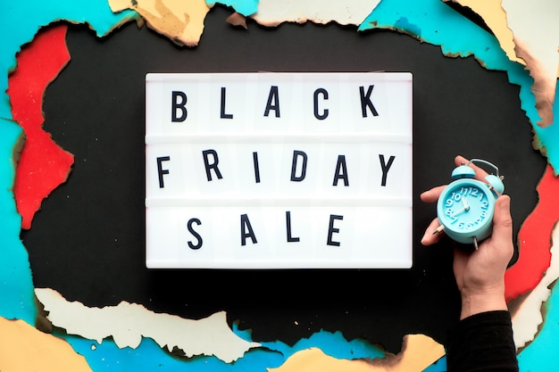 Lightbox-Text "Black Friday Sale" in gebranntem Papierloch in weißem, rotem, gelbem und türkisfarbenem Papier mit verbrannten Kanten, kreative Wand aus schwarzem Papier, Hand in schwarzem Hemd mit Wecker.