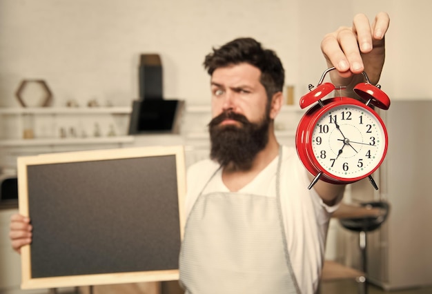 Foto ligue para o tempo de entrega para refeição comida saudável e bebidas governanta masculina na cozinha cozinhando por receita hipster barbudo oferecer menu foco seletivo cópia espaço homem com placa e despertador