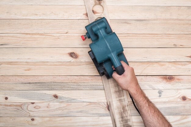 Lijadora eléctrica de banda, lijadora en mano masculina. Procesamiento de pieza sobre mesa de madera marrón claro.