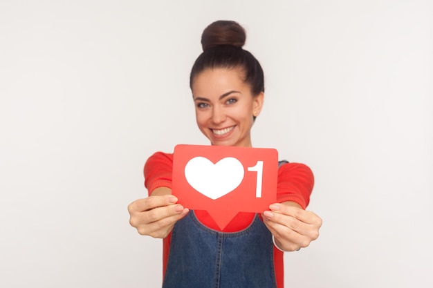 Like und folge interessanten Inhalten. Porträt eines charmanten, positiven, trendigen Mädchens mit Haarknoten in Denim-Overalls, das der Kamera ein Herzsymbol für soziale Medien zeigt und lächelt. Studioaufnahme im Innenbereich, isoliert