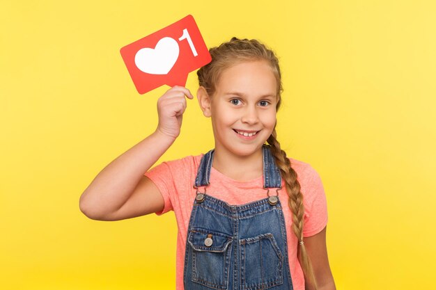 Liken und kommentieren Sie das Bloggen von Kindern. Porträt eines glücklichen kleinen Mädchens in Denim-Overalls mit Herzknopf für soziale Netzwerke, Follower-Benachrichtigungssymbol, beliebte Internetinhalte. Studioaufnahme isoliert