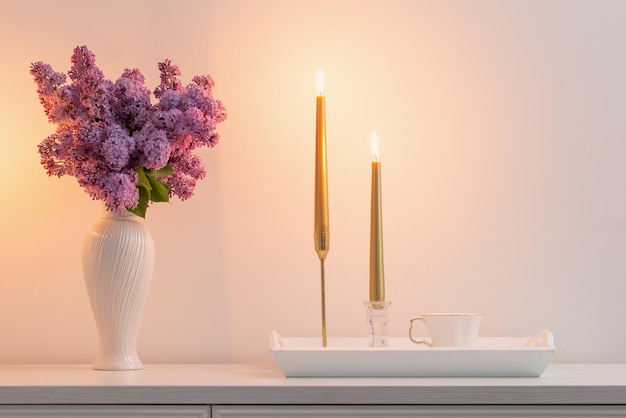 Lila Blumen in weißer Vase und goldene Kerzen auf weißer Hintergrundwand